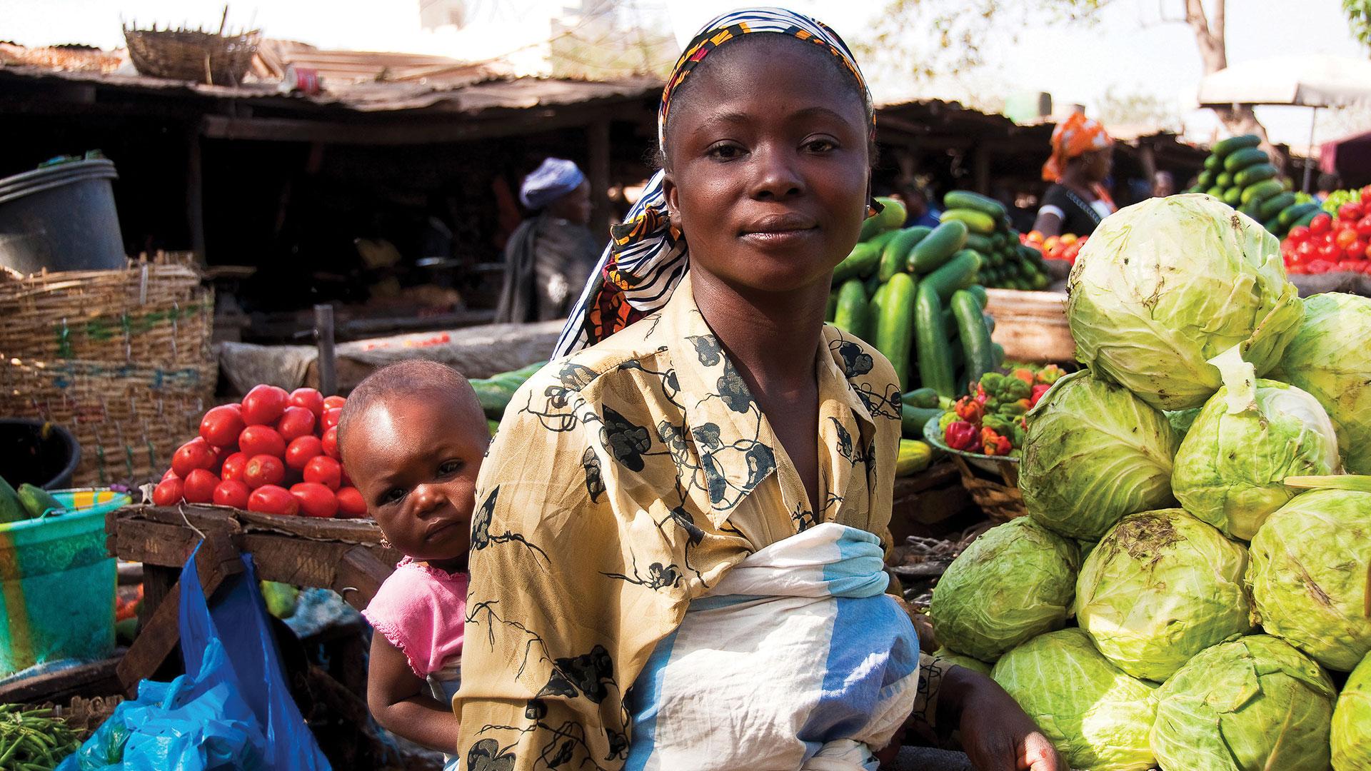 Vegetable stand owner carries her daughter on her back at an open market in Bamako, Mali (Photo: Bigstock)